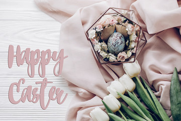 happy easter text greeting card sign on stylish easter egg with floral ornaments in nest of flowers and tulips on rustic white wooden background flat lay. happy easter. space for text, top view