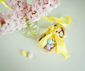 Treat candy for Easter in basket with an yellow bow and glass vase with hyacinths on white table. Empty copy space for text