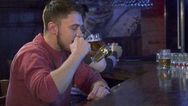 Young caucasian man tasting beer at the pub. Attractive bearded guy getting his lagger from bartender's hand. Side view of male customer putting the glass with beverage on the bar counter