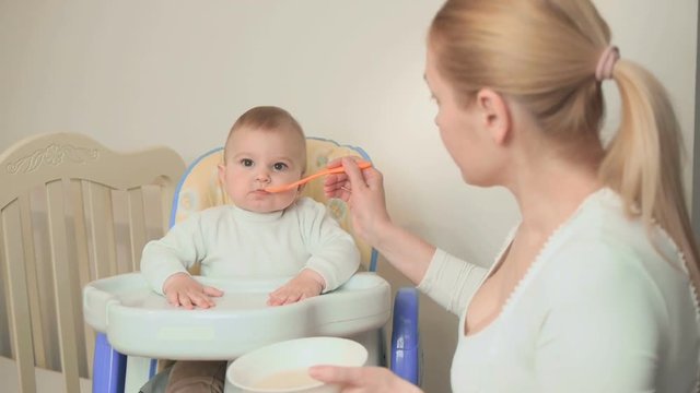 Mother tries to feed a small playful and cheerful child with vegetable puree in the kitchen of the house