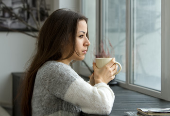 Girl with a cup. / A girl in a bright sweater holding a cup near the lips and looking into the distance through the window, leaning on the wooden sill.