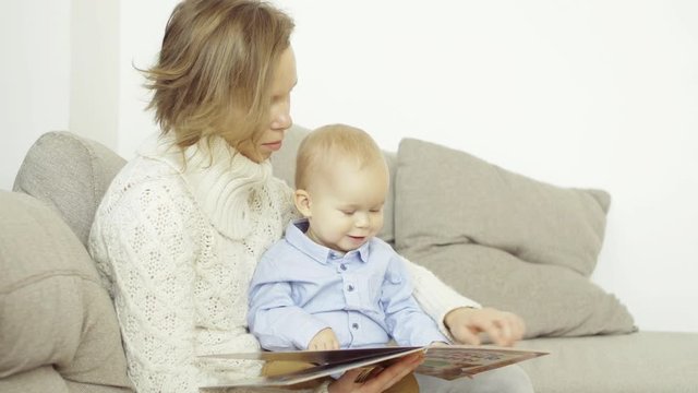 Caring mother reads a book and shows pictures to her little baby, sitting on the sofa at home, son sits on his mother's lap