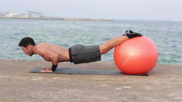 Athletischer Mann macht Liegestuetze mit Fitnessball am Meer
