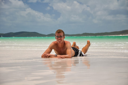 Young man in board shorts wearing sunglasses lying on the white sand of Whitehaven Beach along Whitsunday Island, Queensland, Australia. Whitehaven beach is a popular tourist destination. Clear water.