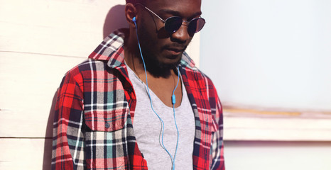 Fashion portrait young african man listens to music in headphones, hipster wearing a plaid red shirt and sunglasses over city background