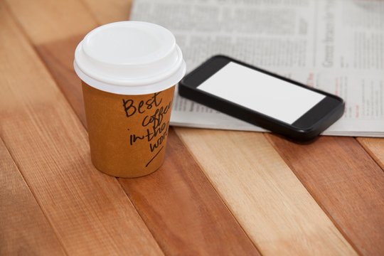 Close-up of disposable coffee cup, mobile phone and newspaper
