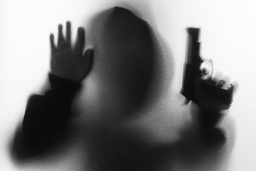 Shadow blur of horror man in jacket with hood gun in his hand.Dangerous man behind the frosted glass.Mystery man.Halloween background.Black and white picture.Blur picture.