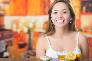 Young brunette sitting by table with glass of honey, granola and sliced lemon in front, smiling happily