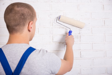 back view of man painter in workwear painting brick wall with paint roller