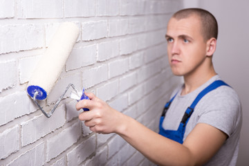 young handsome man painter in workwear painting brick wall with paint roller