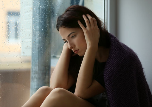 Depressed young woman sitting near window at home
