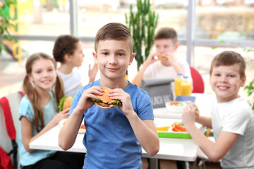 Cute boy eating hamburger and children sitting at table in school cafeteria