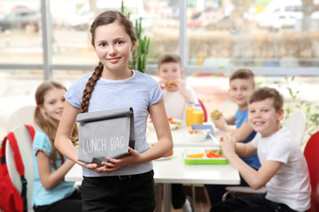 Cute girl holding lunch bag and children eating at table in school cafeteria