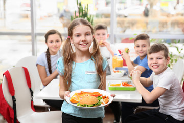 Cute girl holding plate with delicious food and children eating at table in school cafeteria