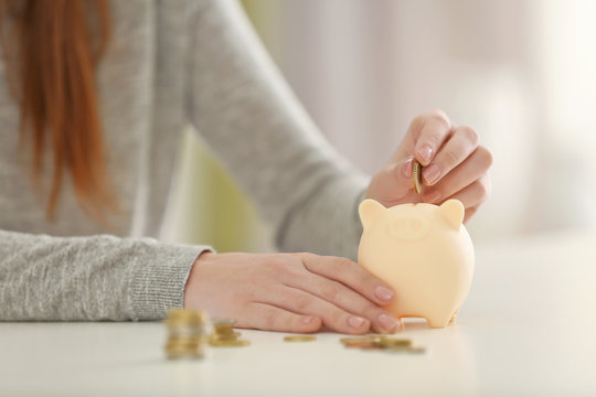 Young woman putting coins into piggy bank at home, closeup