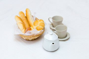 Two cups on saucers teapot wicker straw basket with bread light breakfast snack on white background