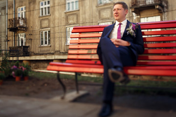 Handsome blond groom with violet boutonniere sits on red bench