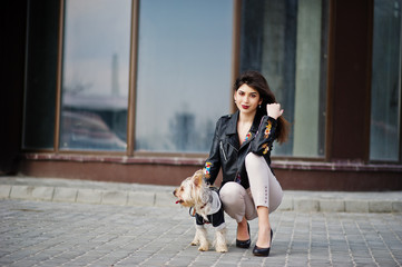 Brunette gypsy girl with yorkshire terrier dog posed against large windows house. Model wear on leather jacket and t-shirt with ornament, pants and shoes with high heels.