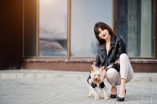 Brunette gypsy girl with yorkshire terrier dog posed against large windows house. Model wear on leather jacket and t-shirt with ornament, pants and shoes with high heels.