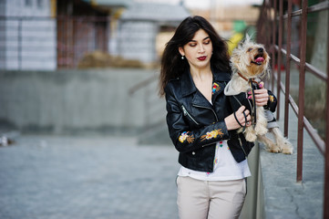 Brunette gypsy girl with yorkshire terrier dog posed against steel railings. Model wear on leather jacket and t-shirt with ornament, pants.