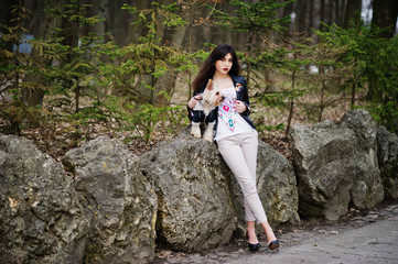 Naklejka na ściany i meble Brunette gypsy girl with yorkshire terrier dog posed against stones on park. Model wear on leather jacket and t-shirt with ornament, pants and shoes with high heels.
