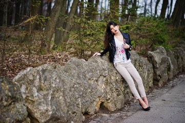 Naklejka na ściany i meble Brunette gypsy girl posed against stones on park. Model wear on leather jacket and t-shirt with ornament, pants and shoes with high heels.