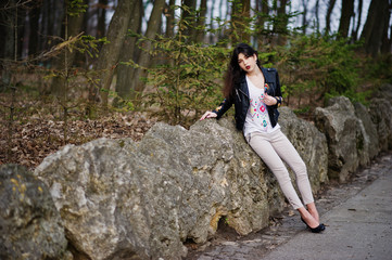 Naklejka na ściany i meble Brunette gypsy girl posed against stones on park. Model wear on leather jacket and t-shirt with ornament, pants and shoes with high heels.