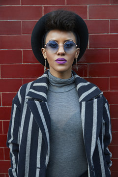 Young woman in hat and sunglasses against a wall, waist up