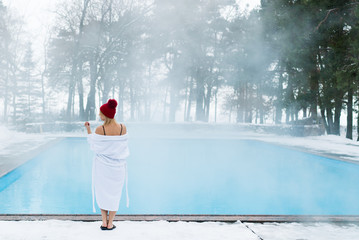 Young blonde woman in bathrobe and red hut near outdoor swimming pool at winter day