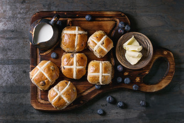 Hot cross buns on wooden cutting board served with butter, knife, fresh blueberries and jug of...