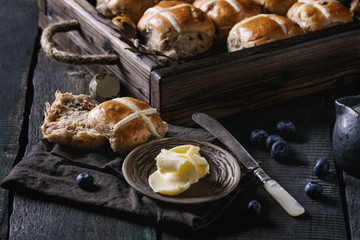 Hot cross buns in wooden tray served with butter, knife, blueberries, easter eggs, birch branch, jug of cream on textile papkin over old texture wood background. Dark rustic style. Easter baking.