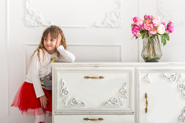 The little girl in a red skirt leaned on the dresser. On the dresser is a bouquet of tulips