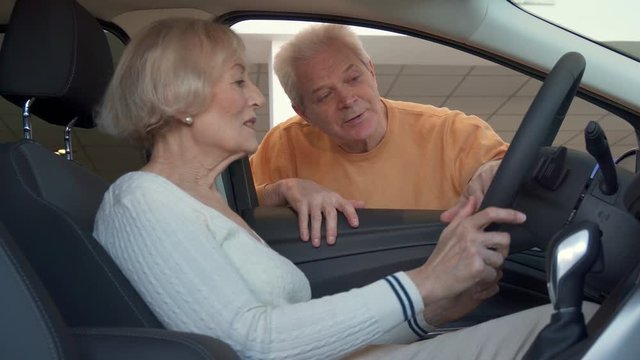 Senior caucasian woman sitting inside the car at the dealership. Aged blond woman keeping her hands on the steering wheel. Attractive gray man explaining something on dashboard of the vehicle to his
