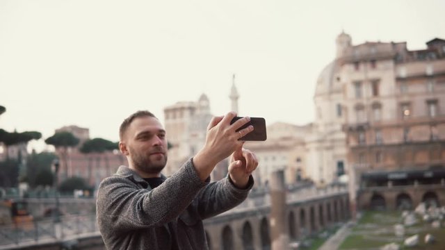 Tourist man takes selfie photos against the background of city centre of Rome, Italy with his smartphone, smiling.