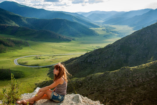 The girl at the cliff on the mountain. Tourist in shorts sitting near the edge of the cliff. At the bottom of a beautiful valley with green hills and mountains and winding road. Mountain Altai, Russia