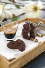 rustic cookies with cocoa and pistachio nuts on wooden tray, single and stacked tied with cloth ribbon, cup of espresso coffee, cloth tablecloth and white flowers in the background