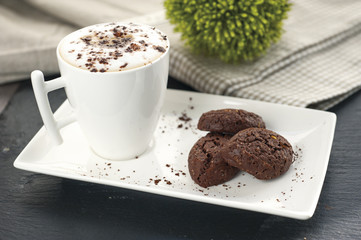 ustic cookies with cocoa and pistachios on white tray, ceramic foam cup with cappuccino and cocoa powder, cloth tablecloth and other kitchen items in the background