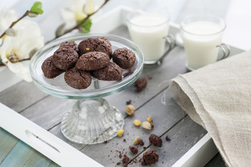 rustic cookies with cocoa and pistachio nuts on cake-stand for sweets with wooden tray two cups milk, cloth napkin and white flowers in the background