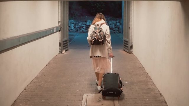 Young woman walking through tunnel carrying the suitcase. Girl with backpack walking the city, traveling. Slow motion.