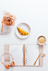 Desktop flatlay hero image, with pink planner, pen, croissant, headphones and rose gold stationery accessories, on a white background. Items arranged to the bottom with negative space at the top