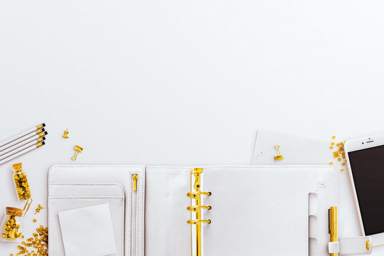 Open Planner Flatlay On A White Desk Background, With White And Gold IPhone And Gold Stationery Accessories. Negative Space Above.