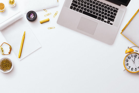 Desktop flatlay scene, with Macbook laptop, notepad, pens along with other gold stationery items, on a plain white desk background. Negative space below