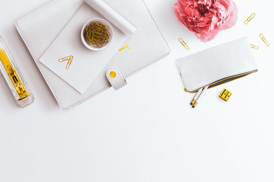 A feminine desktop flatlay hero image, with a closed planner, pink peony and gold stationery accessories. Negative space to the centre bottom, on a plain white desk background.