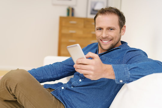 glücklicher mann sitzt entspannt auf dem sofa und hält sein mobiltelefon in der hand