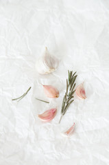 The head of garlic and garlic cloves with rosemary lie on the background of white crumpled paper. Vertical image of food, daylight.