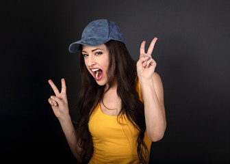 Obraz na płótnie Canvas Young crazy happy female model in blue hat showing victory gesture on grey dark background with empty copy space.