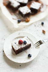 Table setting, chocolate dessert brownie cake with berries on a white plate on a table covered with a linen tablecloth. The second part of the cake on the parchment paper. Vertical image, daylight.