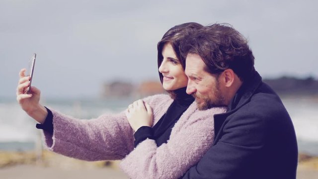 Very happy couple in love taking selfie on the beach in winter slow motion making funny faces