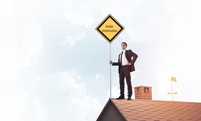 Young businessman on house brick roof holding yellow signboard. 