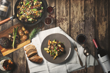 Overhead view of colorful roast vegetables, savory sauces and salt served with grilled t-bone steak on a rustic wooden counter in a country steakhouse